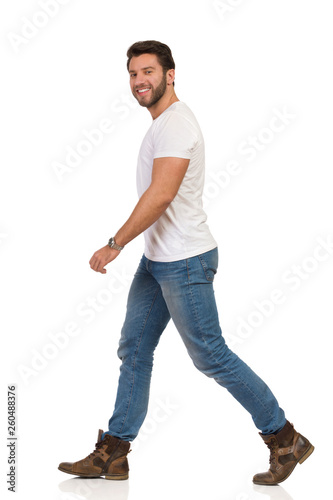 Young Man In Jeans And White T-shirt Is Walking And Looking At Camera. Side View.