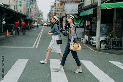 two happy female friends travelers with bags crossing street together outdoor sunny day in china town. japanese lady travel in chinese city walking on zebra cross in urban. girl wear hats look sky.