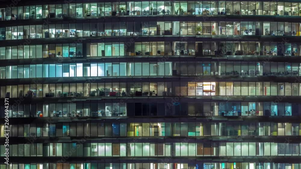 Working evening in glass office building with numerous offices with glass walls and illuminated windows timelapse. People sitting at desks. Pan left Working evening in glass office building with numerous offices with glass walls and illuminated windows timelapse. People sitting at desks. Pan left