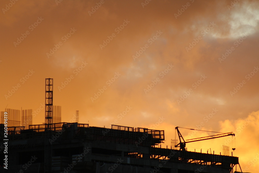 Silhueta de guindaste e edifício em construção no pôr-do-sol com o céu repleto de nuvens na cor laranja