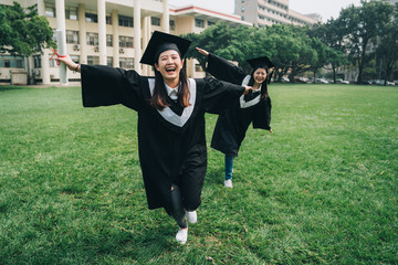 full length two young positive students graduation ceremony concept. beautiful elegant smiling college girls in gowns and mortarboard running joyful on lawn outdoor spring park in university campus