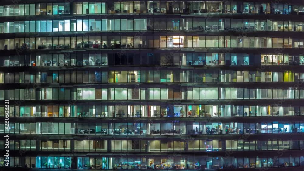 Working evening in glass office building with numerous offices with glass walls and illuminated windows timelapse. People sitting at desks. Pan right Working evening in glass office building with numerous offices with glass walls and illuminated windows timelapse. People sitting at desks. Pan right