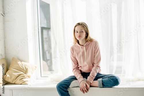 Pensive teenage kid in jeans sitting on window sill and looking away