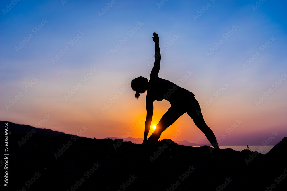 Silhouette of a young woman doing yoga on the beach at sunset.