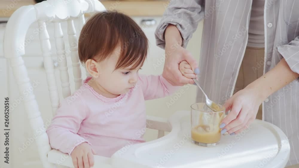 Unrecognizable mother helping her baby daughter eat fruit puree with spoon in highchair Unrecognizable mother helping her baby daughter eat fruit puree with spoon in highchair