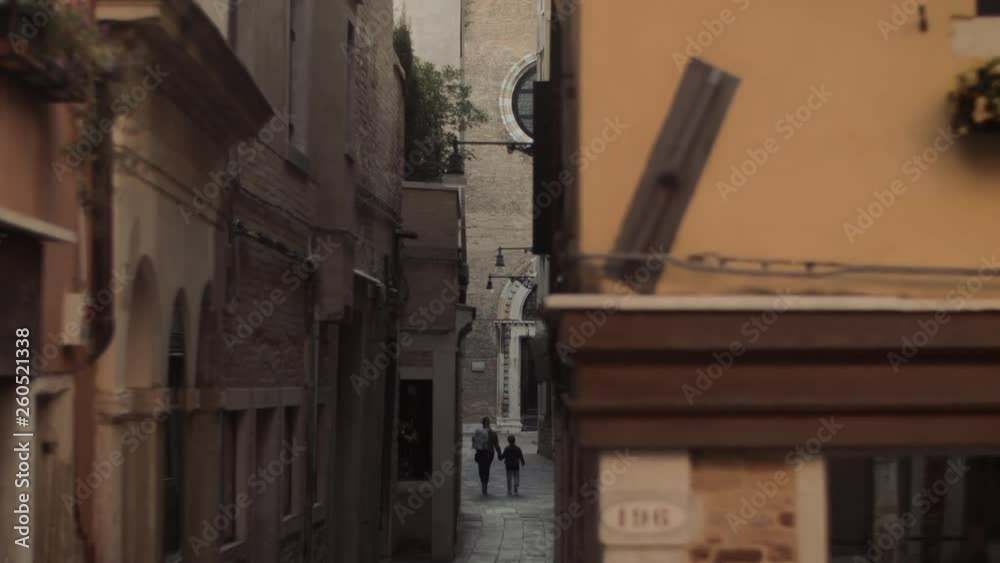 A beautiful narrow Venice street. Ancient stone facades, paved cover, wall mounted streetlights. A woman and a girl are walking down it holding hands A beautiful narrow Venice street. Ancient stone facades, paved cover, wall mounted streetlights. A woman and a girl are walking down it holding hands