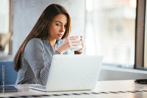 Manager drinking coffee at the office, sitting in front of her laptop. 