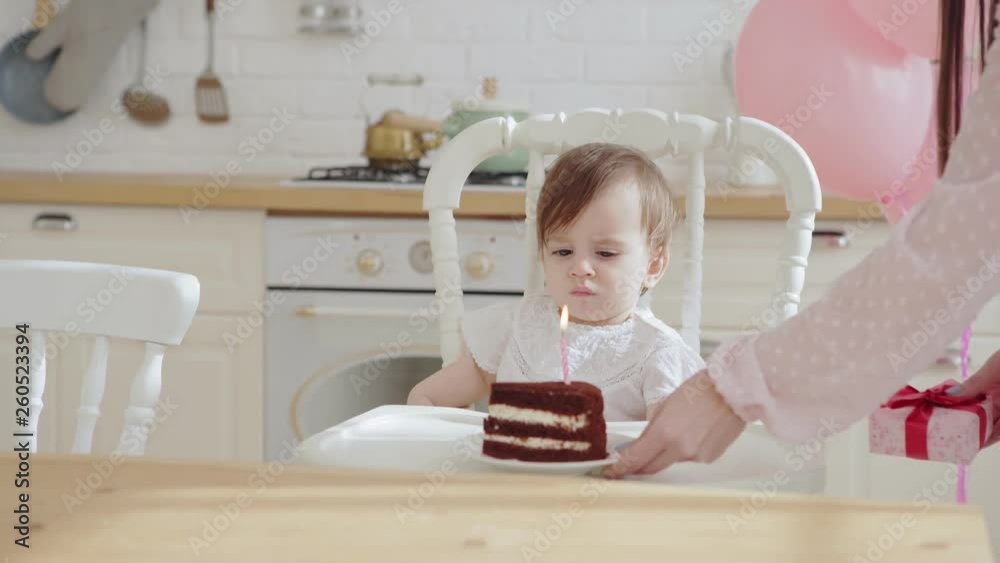 Loving mother giving baby daughter sitting in highchair a piece of birthday cake with one burning candle on her first birthday celebration Loving mother giving baby daughter sitting in highchair a piece of birthday cake with one burning candle on her first birthday celebration