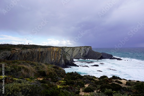 Dark, purple clouds over the sea, storm, large waves, cliffs, lighthouse just visible in the distance. Portugal 