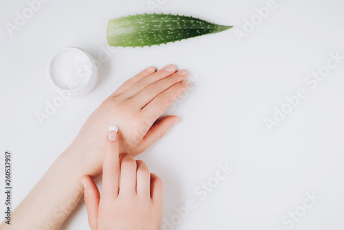 Closeup of beautiful female hands applying cream. Organic cream from Aloe vera fresh leaves on white background. Flat lay, top view, copy space. Healthcare concept.