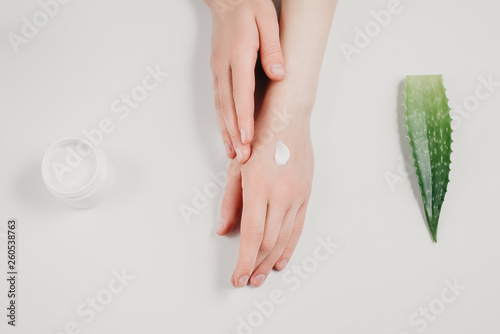 Beautiful groomed woman s hands with organic cream jar and Aloe vera fresh leaves on white background. Moisturizing cream for clean and soft skin. Flat lay  top view  copy space. Healthcare concept.