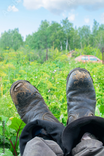 Legs in shabby work boots of man that relax at garden with green forest background