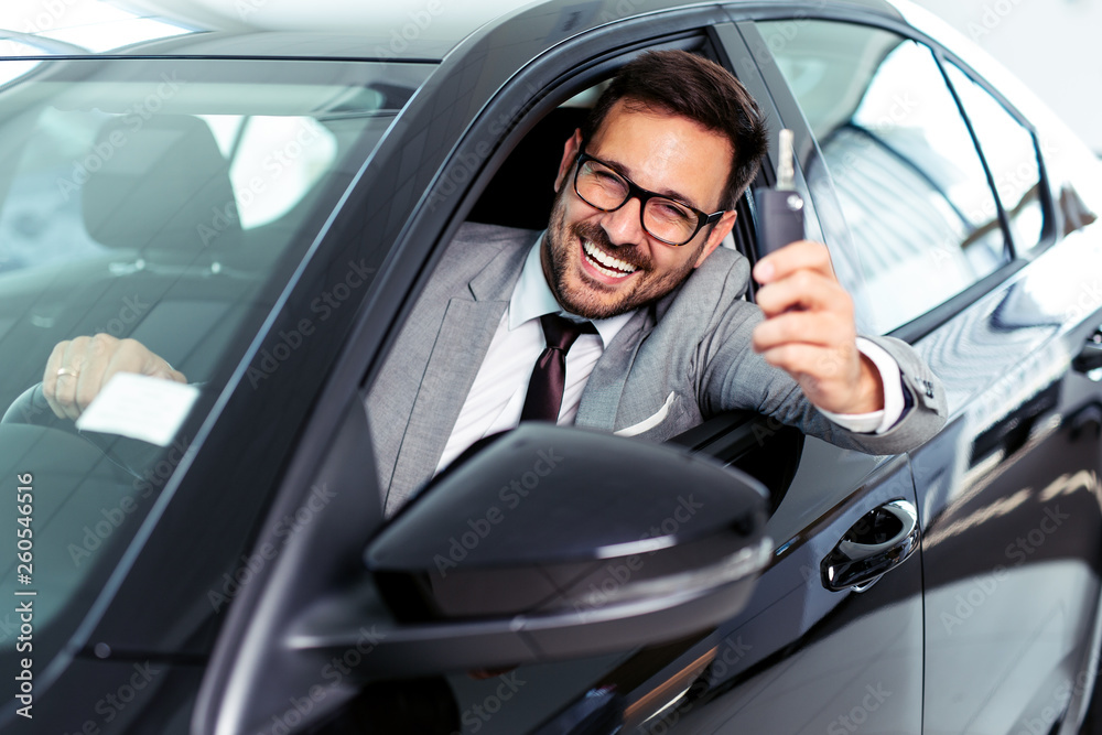 Young businessman sitting in car and showing car keys in car dealership showroom