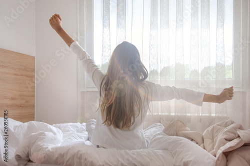 Fotografia Young woman waking up in her bedroom, sitting on the bed stretching arms by the