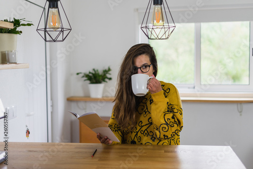 Mulher de óculos bebendo uma bebida numa caneca branca enquanto segura um caderno aberto na outra mão, numa bancada de madeira