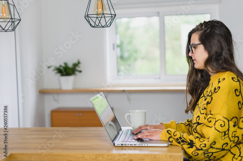 Mulher de óculos a trabalhar no seu computador portátil vista de lado. Caneca branca do seu lado direito.
