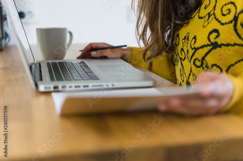 Mulher a trabalhar no seu computador portátil vista de lado. A segurar um caderno de apontamentos e com uma caneca branca do seu lado direito.