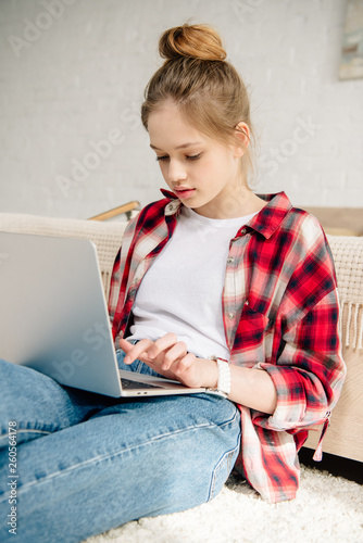 Teenage kid in checkered shirt using laptop while sitting on carpet