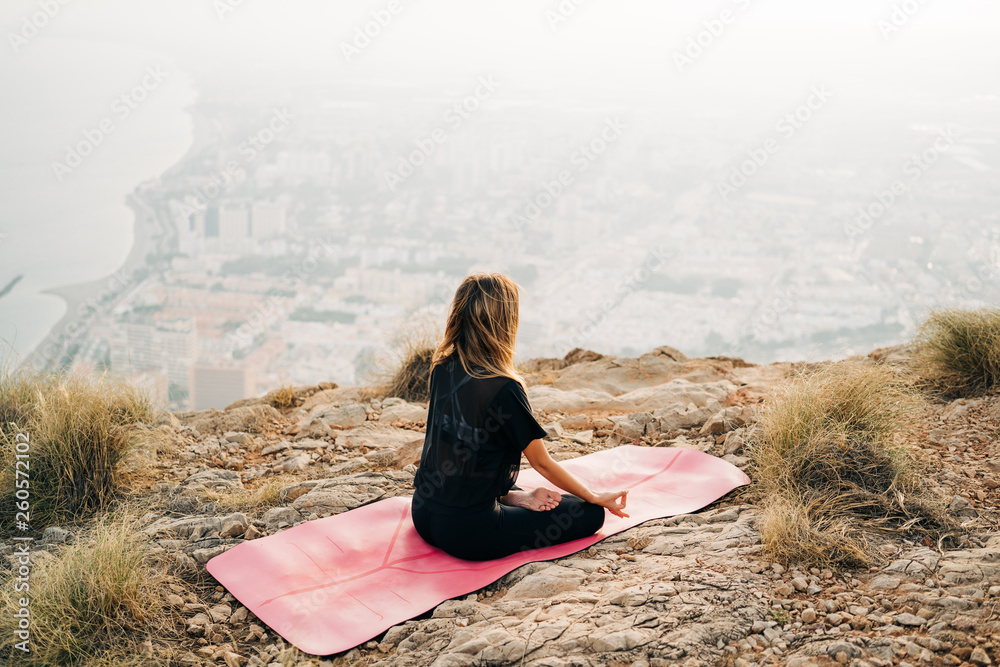 Beautiful young woman doing yoga at the top of the mountain with views from the city during a nice sunset. Freedom and yoga concept.