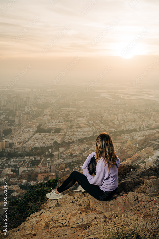 Young woman sitting and using the mobile on the top of the mountain looking at the city from above, during nice sunset
