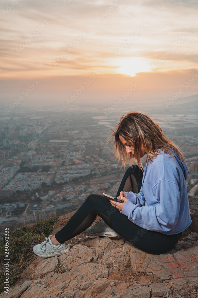 Young woman sitting and using the mobile on the top of the mountain looking at the city from above, during nice sunset Young woman sitting and using the mobile on the top of the mountain looking at the city from above, during nice sunset