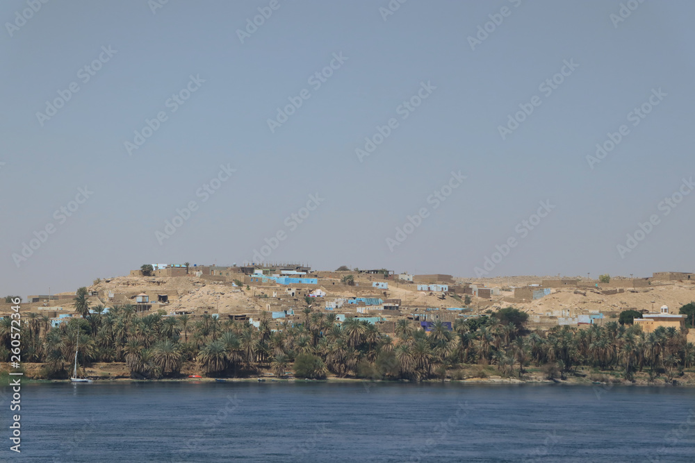 Blick auf ein typisches Dorf in Ägypten nahe Luxor am Ufer des Nil, blaues Wasser, blauer Himmel, Wüste