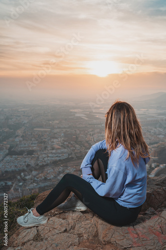 Young woman sitting and using the mobile on the top of the mountain looking at the city from above, during nice sunset © ManuPadilla