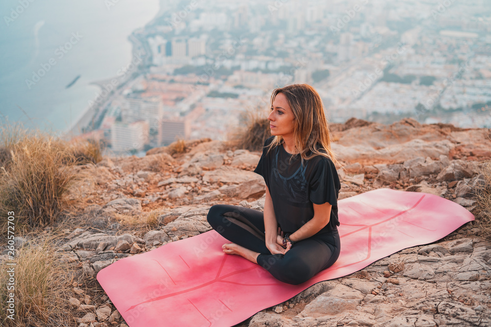Beautiful young woman doing yoga at the top of the mountain with views from the city during a nice sunset. Freedom and yoga concept.