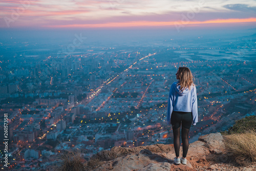 Young woman at the top of the mountain looking and points the city from above, during nice sunset