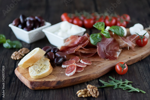 Prosciutto, bread, olives, walnut, mozzarella, salami, basil and cherry tomatoes on brown wooden board. Mediterranean kitchen.