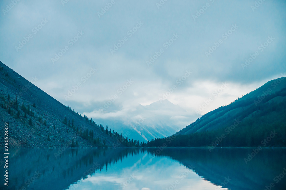 Amazing silhouettes of mountains and low clouds reflected on mountain lake. Beautiful ripples on water mirror. Cloudy sky in highlands. Atmospheric ghostly landscape. Wonderful mystic mountainscape.