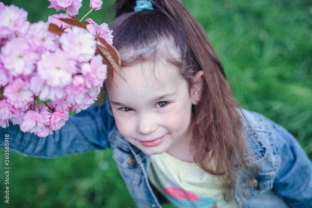 Adorable little girl with brown hair in blue denim jacket having fun in blossom cherry garden on beautiful spring day. Adorable little girl with brown hair in blue denim jacket having fun in blossom cherry garden on beautiful spring day.
