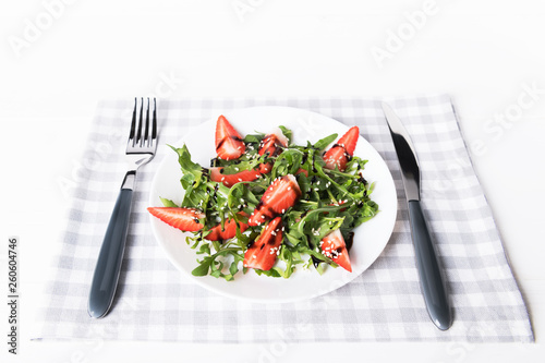 Healthy organic diet salad with arugula, strawberries and sesame in white plate on grey napkin on a white wooden background