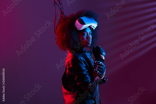 Stylish curly dark haired girl dressed in black leather jacket and gloves is wearing the virtual reality glasses on her head in the dark studio with neon light