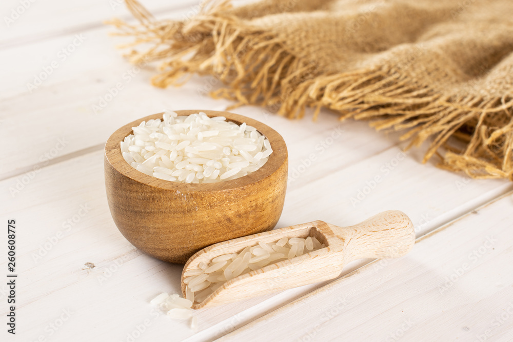 Lot of whole white jasmine rice grains in a wooden bowl with wooden scoop on jute cloth on white wood Lot of whole white jasmine rice grains in a wooden bowl with wooden scoop on jute cloth on white wood
