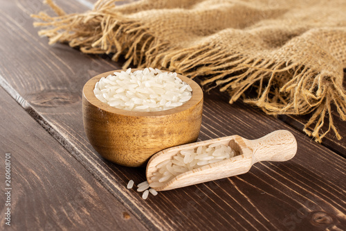 Lot of whole white jasmine rice grains in a wooden bowl with wooden scoop on jute cloth on brown wood