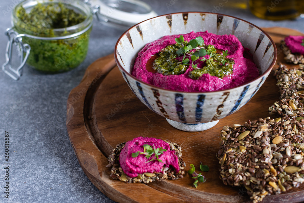 Roasted beet hummus in ceramic bowl, flax seed crackers on wooden cutting board, pesto sauce jar - vegetarian, vegan food concept Roasted beet hummus in ceramic bowl, flax seed crackers on wooden cutting board, pesto sauce jar - vegetarian, vegan food concept