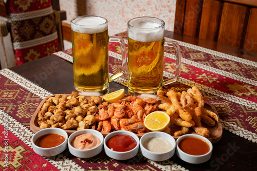 Beer set.Two Beers and appetizers on a wooden plate with five sauces.On a wooden table with a tablecloth with national patterns.