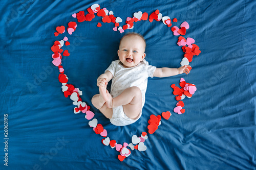 Cute smiling white Caucasian baby girl boy infant with blue eyes four months old lying on bed among many foam paper red pink colorful hearts. View from top above. Happy Valentine day
