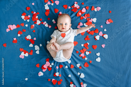 Cute adorable white Caucasian baby girl boy infant with blue eyes four months old lying on bed among many foam paper red pink colorful hearts. View from top above. Happy Valentine day holiday