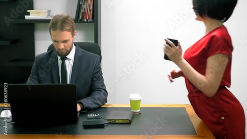 Office flirt. Attractive woman in red overalls with a deep neckline flirting at the table with her colleague. man looks at the chest of a woman in the office