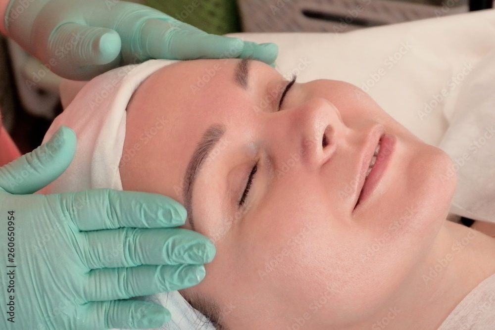Smiling sweet girl lying in a chair at the beautician. The doctor specialist in gloves rubs the cream in the woman's face. Treatments at the beauty salon Smiling sweet girl lying in a chair at the beautician. The doctor specialist in gloves rubs the cream in the woman's face. Treatments at the beauty salon