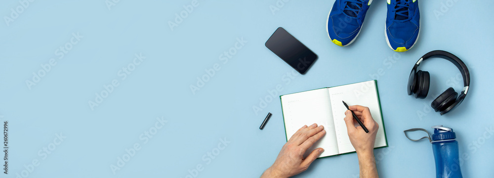 Male hands writing in notebook diary, headphones, blue sneakers, bottle of waters, smartphone on blue background Top view flat lay copy space. Concept of active lifestyle sports fitness health - obrazy, fototapety, plakaty 