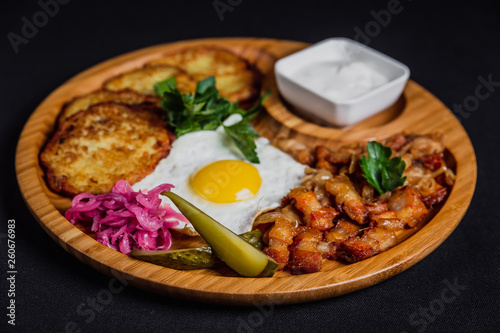 Potato pancakes with a fried egg, bacon, sourcream, horse radish, pickles and parsley on a wooden plate on black background