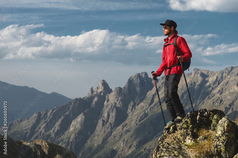Porter of a bearded traveler hipster in sunglasses and a cap with a backpack and trekking poles stands on a high cliff near the cliff on the background of epic mountains of rocks and clouds