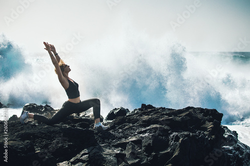 The girl practices yoga on the ocean. Waves are breaking on stones. Beautiful background.