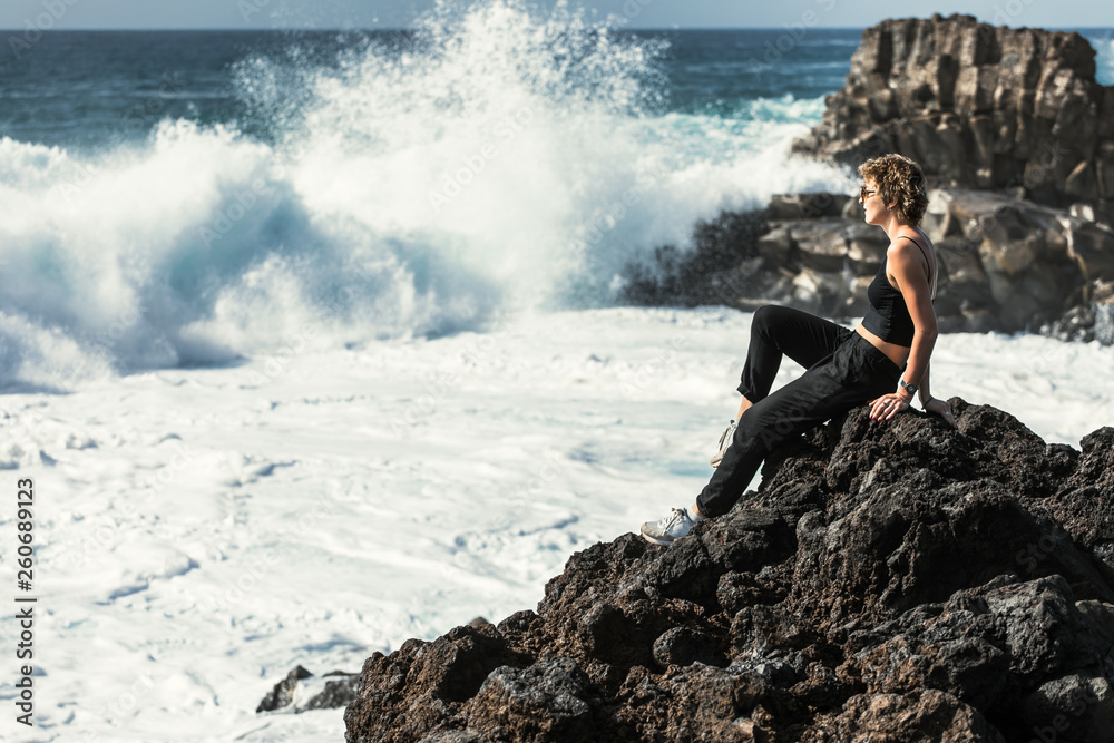 The girl is sitting by the ocean. Huge waves are breaking on the rocks. Volcanic beach. Tenerife Spain.