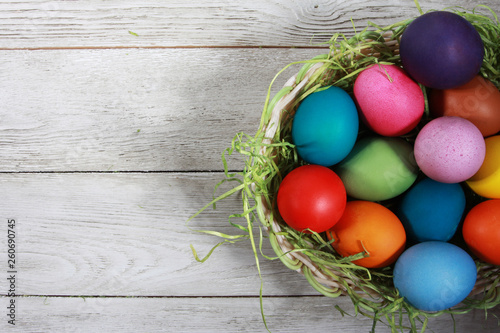 Colorful Easter eggs and pile of green grass in basket on white wooden table. Easter background, egg hunt concept. Blank copy space, mockup, hay