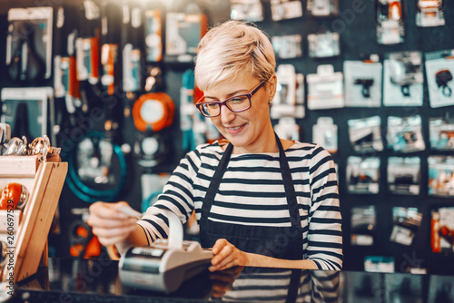 Smiling Caucasian female worker with short blonde hair and eyeglasses using cash register while standing in bicycle store. photo
