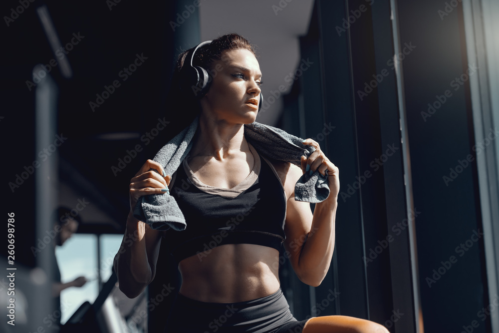 Fototapeta premium Young attractive strong muscular female bodybuilder with ponytail and headphones posing in gym with towel around neck. When life is tough, remember you are tougher.